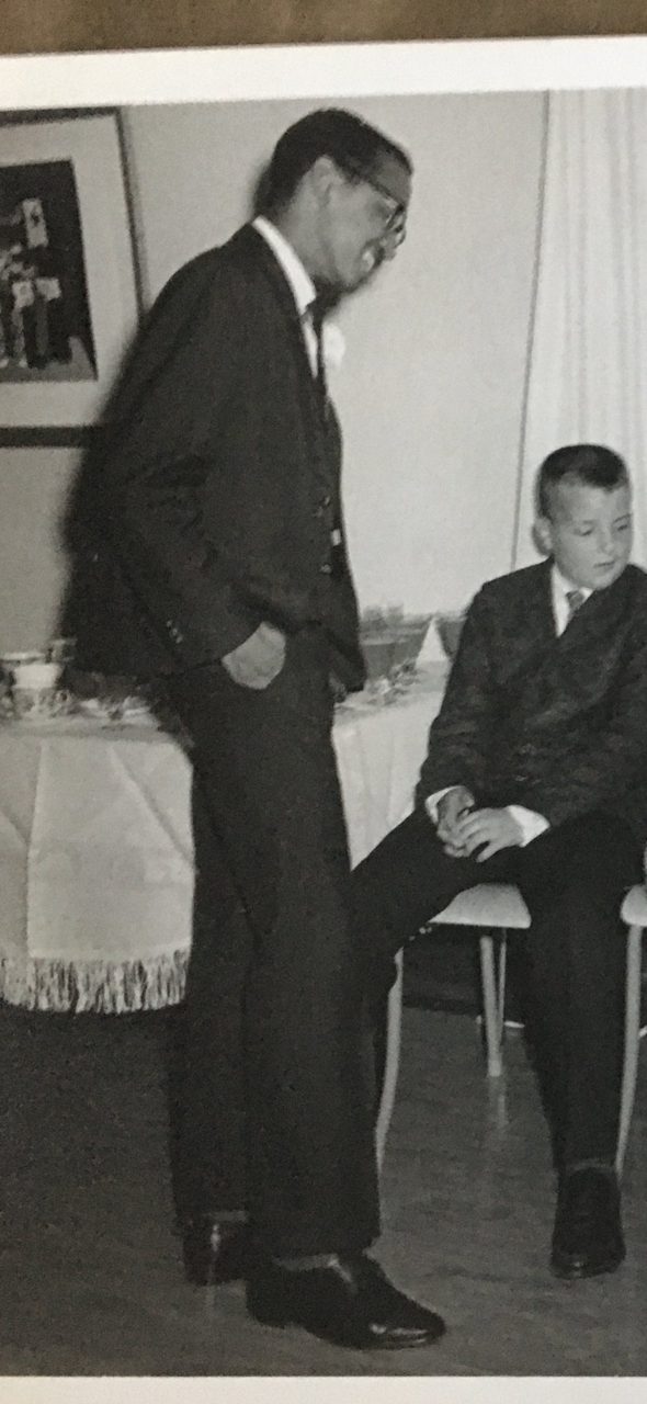 A black-and-white photograph of young Gabriel in a suit and glasses, laughing, standing in a living room in Stratford
